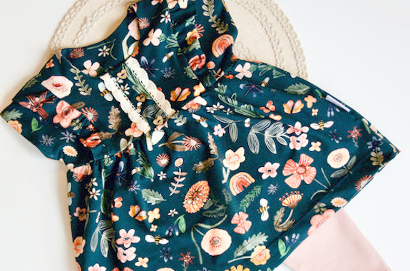 Flatlay einer Tunika und Leggings. Die Tunika ist aus dunkelblauem Biojersey mit verschiedenen Blumen in rosa, ocker und grün. Auch Regenbögen und Bienchen sind drauf. Die Leggings ist Rosa. Biostoffe bei Faybee.ch kaufen