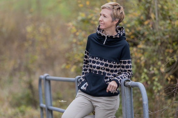 Frau steht im Freien. Sie trägt einen selbstgenähten Hoodie mit sportlicher Teilung und Reissverschluss am Kragen. Stoffe sind von fayee.ch